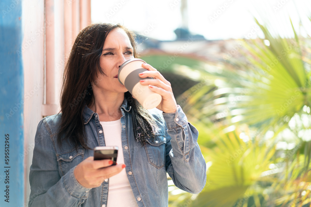Middle aged woman using mobile phone and holding a coffee
