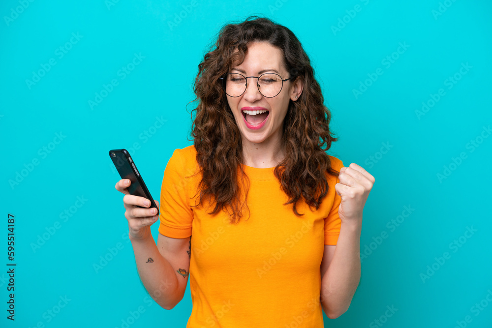 Young caucasian woman isolated on blue background using mobile phone and doing victory gesture Young caucasian woman isolated on blue background using mobile phone and doing victory gesture