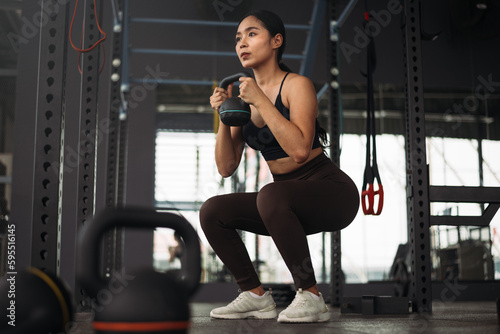 Strong asian woman doing exercise with kettlebell at crossfit gym. Athlete female wearing sportswear workout on grey gym background with weight and dumbbell equipment. Healthy lifestyle.