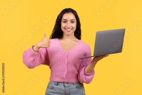 Glad millennial arab female in casual work on laptop, show thumb up isolated on yellow background