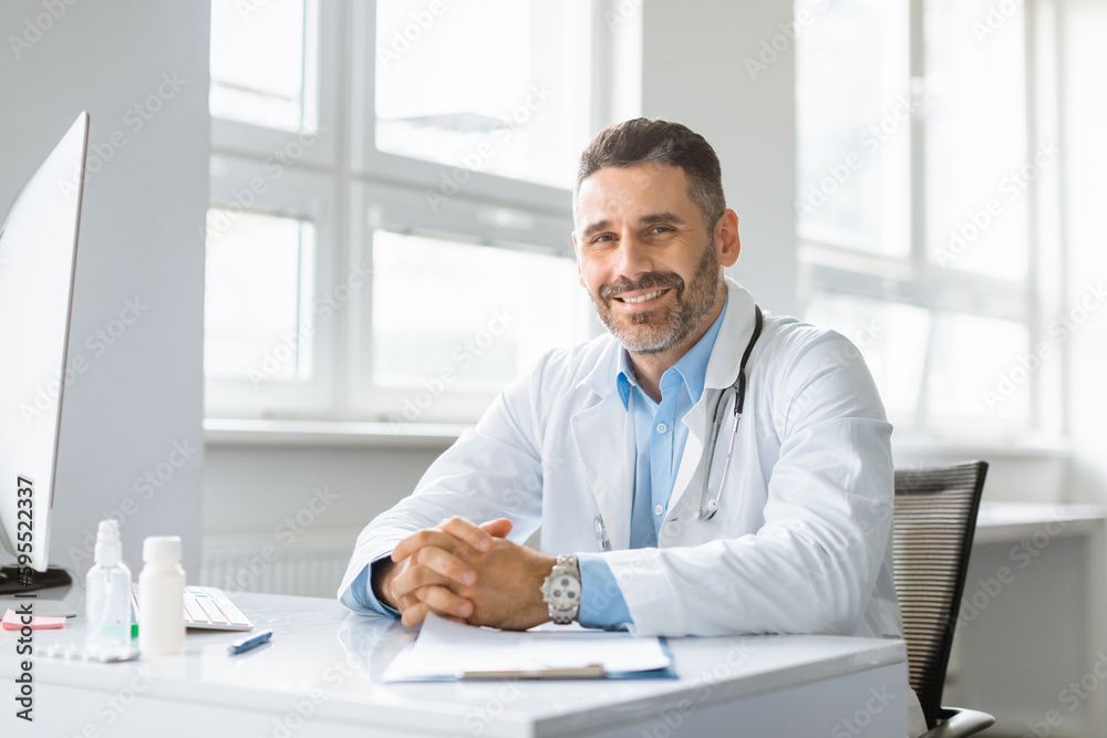 Portrait of happy cheerful man in white workwear doctor posing at clinic, sitting at workdesk and smiling at camera