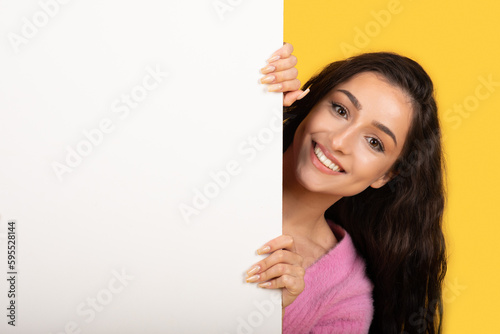 Smiling young arab lady peeks out from behind big banner with empty space for ad, text and offer