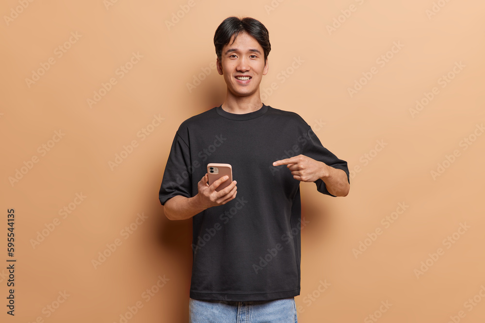 Handsome Chinese man smiles gladfully points at his new smartphone shares awesome news with friends dressed in casual black t shirt isolated over brown background asks to check out newsfeed. Handsome Chinese man smiles gladfully points at his new smartphone shares awesome news with friends dressed in casual black t shirt isolated over brown background asks to check out newsfeed.