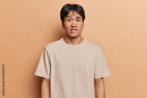 Waist up shot of worried Japanese man bites lips looks anxious and surprisingly at camera doesnt know way out from difficult situation dressed in casual t shirt isolated over brown background