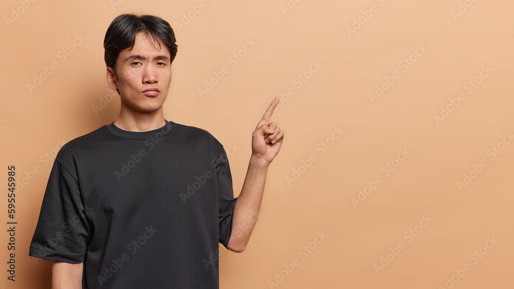 Studio shot of serious self confident Asian man with dark hair dressed in casual black t shirt points index finger at blank space advertises something isolated on brown wall. Place your promo here Studio shot of serious self confident Asian man with dark hair dressed in casual black t shirt points index finger at blank space advertises something isolated on brown wall. Place your promo here