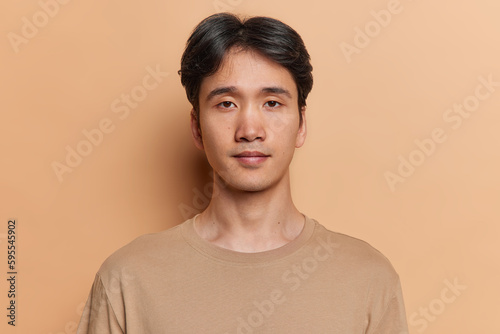 Portrait of serious dark haired adult Asian man with neutral facial expression focused at camera dressed in casual jumper isolated over brown background. Handsome Japanese guy poses in studio photo