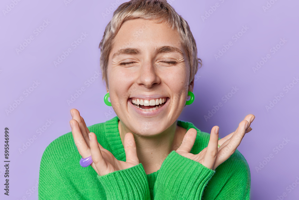 Portrait of short haired woman keeps eyes closed smiles broadly shows white teeth keeps hands near face wears green warm jumper and earrings isolated over purple background. Happy emotions concept Portrait of short haired woman keeps eyes closed smiles broadly shows white teeth keeps hands near face wears green warm jumper and earrings isolated over purple background. Happy emotions concept