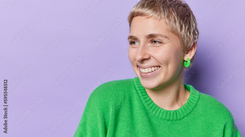 Horizontal shot of positive fair haired woman smiles gladfully focused aside notices something pleasant wears green sweater expresses positive emotions isolated over purple background blank space Horizontal shot of positive fair haired woman smiles gladfully focused aside notices something pleasant wears green sweater expresses positive emotions isolated over purple background blank space