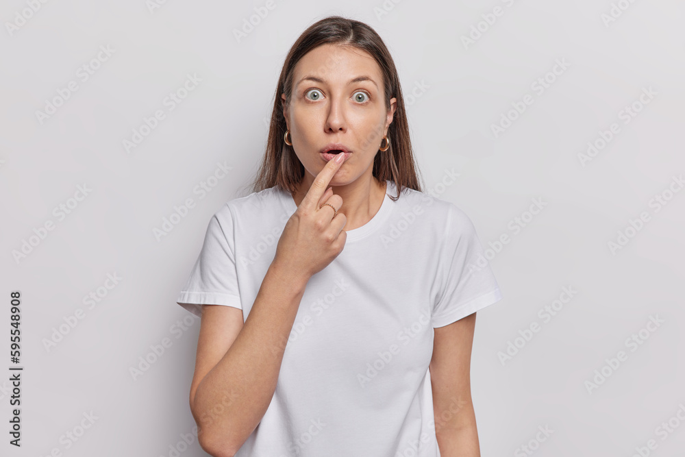 Portrait of stunned woman keeps finger on lips stares bugged eyes feels very shocked dressed in casual t shirt isolated over white background witnesses terrible scene feels scared and intense Portrait of stunned woman keeps finger on lips stares bugged eyes feels very shocked dressed in casual t shirt isolated over white background witnesses terrible scene feels scared and intense