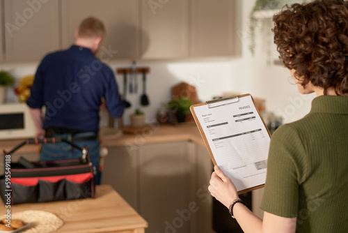 Rear view of young woman examining repair service contract while standing in the kitchen with repairman in background photo