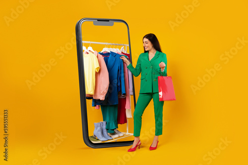 Happy shopaholic woman choosing new outfit on clothing rail in huge cellphone standing on yellow background, full length