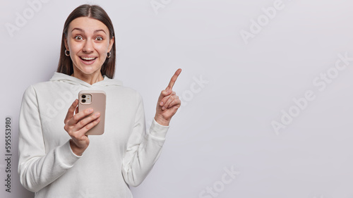 Happy surprised dark haired woman holds smartphone in hand points index finger at copy space gives special offer shows empty mockup place for banner ad wears casual hoodie isolated over white wall