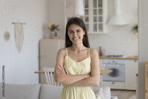 Positive confident pretty brunette young adult Latin girl wearing sleeveless summer dress, looking at camera with crossed hands, smiling, posing at home. Indoor female portrait