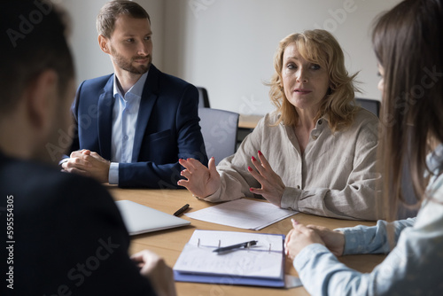 Engaged mature business professional woman talking to colleagues on team meeting, brainstorming, cooperating on project plan, motivating on teamwork, speaking at conference table