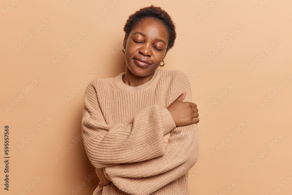 Portrait of dark skinned young woman with short hair embraces herself feels  cozy and comfortable in warm knitted jumper poses against brown background  keeps eyes closed. People self care concept Stock Photo
