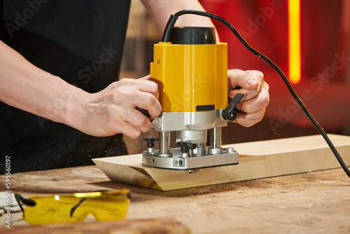 The hands of a caucasian craftsman hold a yellow electric router that processes the edges of a wooden block on a tabletop