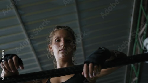 Woman boxer hung from ring ropes in gym after fight. Female muay thai fighter covered in sweat and wearing black sportswear and bandages rests and breathes while looking at camera