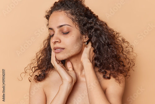 Half naked young woman with natural curly hair touches face gently keeps eyes closed has healthy smooth skin after beauty treatments isolated over brown background. Beauty and wellness concept
