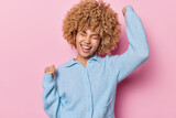 Energetic curly haired woman keeps arms raised up clenches fists exclaims hooray smiles broadly has eyes closed dressed in casual blue jumper isolated over pink background rejoices dream comes true