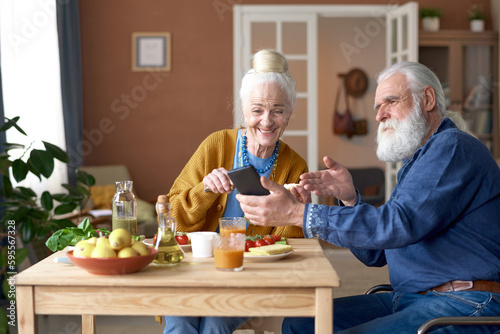 Senior man pointing at screen of smartphone and talking to his wife during breakfast at table at home