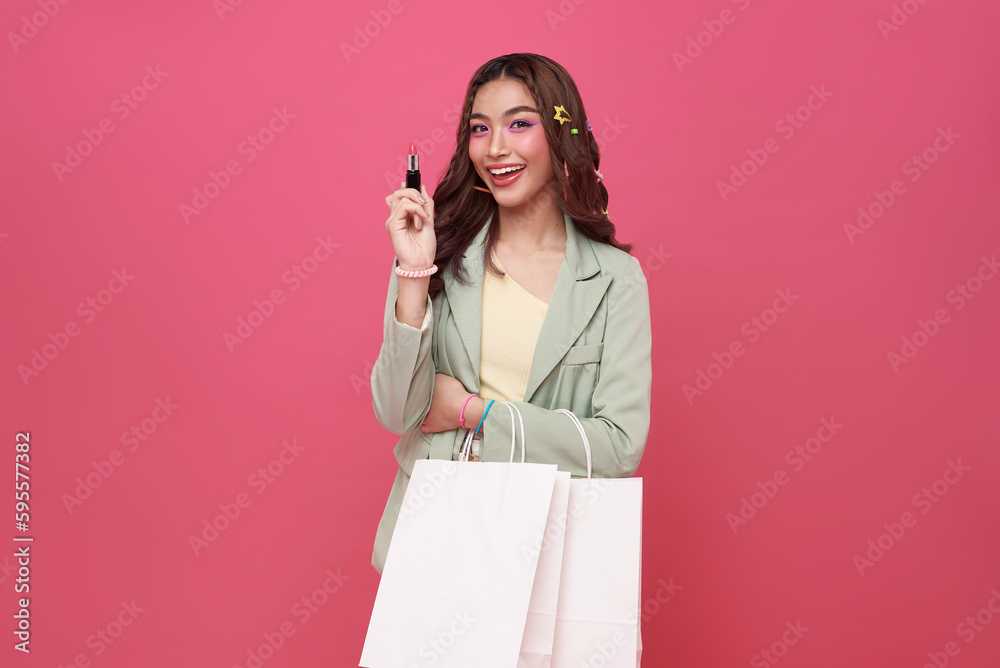 Cheerful happy thai asian woman enjoying shopping, she is carrying shopping bags and showing lipstick isolated on pink background Cheerful happy thai asian woman enjoying shopping, she is carrying shopping bags and showing lipstick isolated on pink background