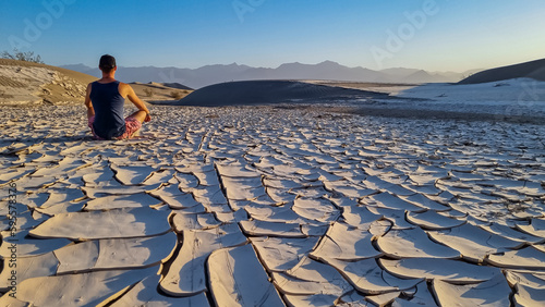 Man sitting on dry cracked clay crust at Mesquite Flat Sand Dunes in Death Valley National Park, California, USA. Looking at pattern Mojave desert in summer with Amargosa Mountain Range in the back photo