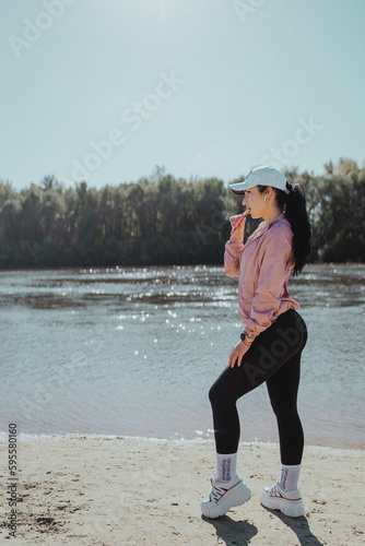 A sports girl in leggings and a cap with a beautiful body on a morning jog on the banks of a river with sand, holding a protein bar in her hand © Alex May