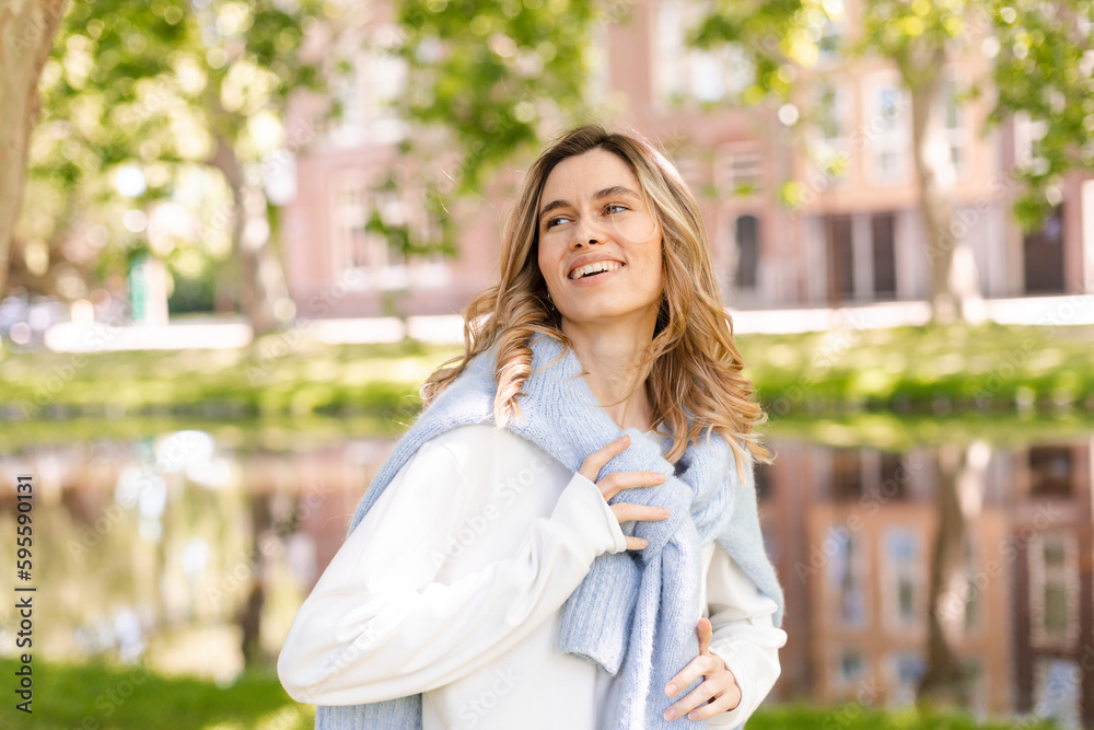 Beautiful woman smiling and look at camera at park at sunny day. Outdoor portrait of a smiling curly blonde girl. Happy cheerful girl laughing at park, look at side, touch sweater, show toothy smile. Beautiful woman smiling and look at camera at park at sunny day. Outdoor portrait of a smiling curly blonde girl. Happy cheerful girl laughing at park, look at side, touch sweater, show toothy smile.