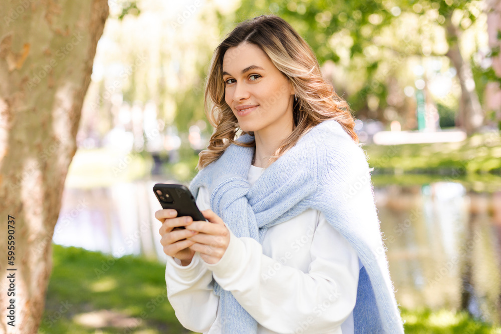 Young stylish curly blonde woman writes message on smartphone walking on park near river, hipster tourist traveling in town and planning trip using mobile phone. Girl chatting on mobile and smiling. Young stylish curly blonde woman writes message on smartphone walking on park near river, hipster tourist traveling in town and planning trip using mobile phone. Girl chatting on mobile and smiling.