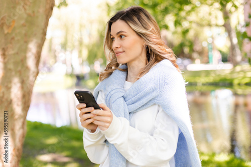 Young stylish curly blonde woman writes message on smartphone walking on park near river  hipster tourist traveling in town and planning trip using mobile phone. Girl chatting on mobile and smiling.
