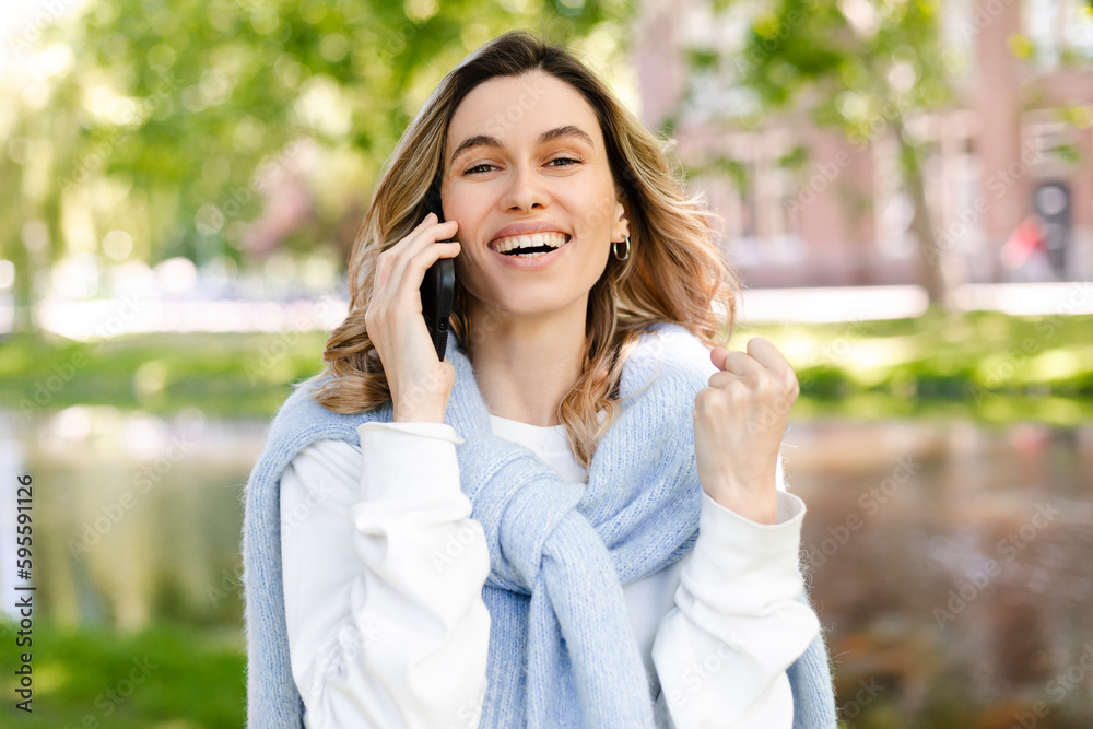 Young happy blonde woman with curly hair received good unexpected news on the phone call in the park. Work promotion, Job offer, great news, girl make win gesture wear blue sweater, white t-shirt. Young happy blonde woman with curly hair received good unexpected news on the phone call in the park. Work promotion, Job offer, great news, girl make win gesture wear blue sweater, white t-shirt.