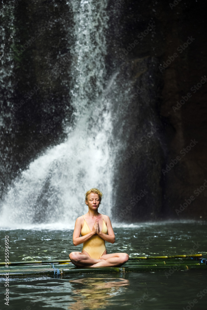 Obraz premium The girl is meditating, her hands are folded in namaste, in the jungle against the backdrop of a waterfall, dressed in a yellow swimsuit.