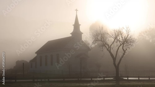 Bright sun pierces heavy fog over small church and tree with bare branches photo