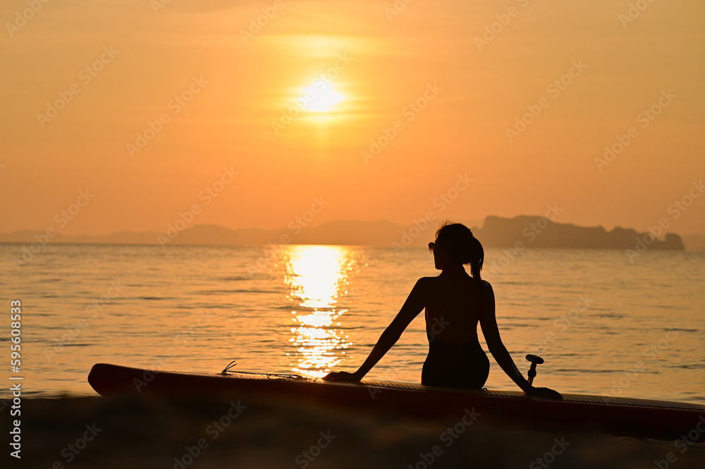 The woman is sitting on the beach with the sup board beside her at sunset.
