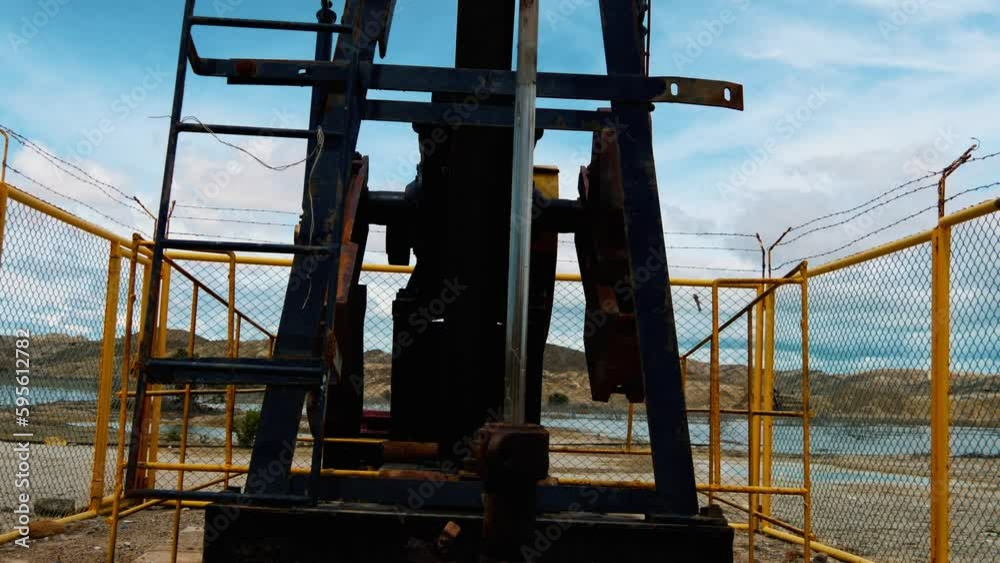 Close-up view of an Oil well pump jack in the Peruvian desert pumping oil next to a small lake of water with cloudy blue skies and desert hills in the background Close-up view of an Oil well pump jack in the Peruvian desert pumping oil next to a small lake of water with cloudy blue skies and desert hills in the background