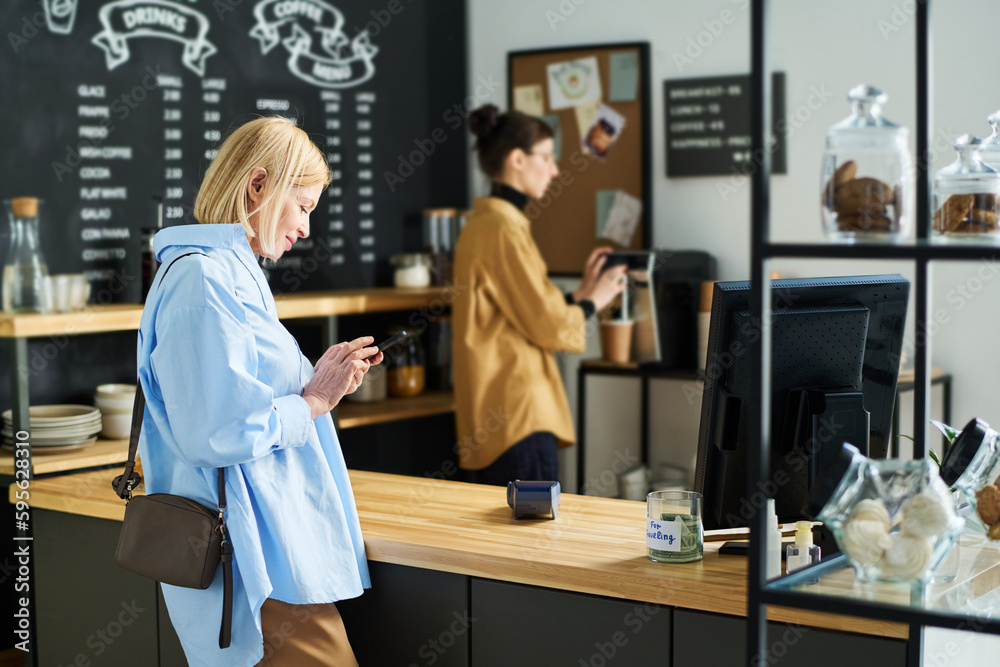 Side view of mature blond woman texting in smartphone while standing by counter desk and waiting for coffee being prepared by clerk Side view of mature blond woman texting in smartphone while standing by counter desk and waiting for coffee being prepared by clerk