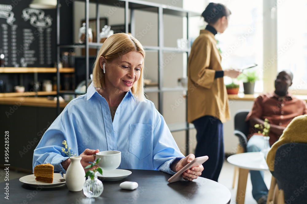 Smiling mature female customer with cup of tea and smartphone watching online video, looking through playlist or communicating in video chat