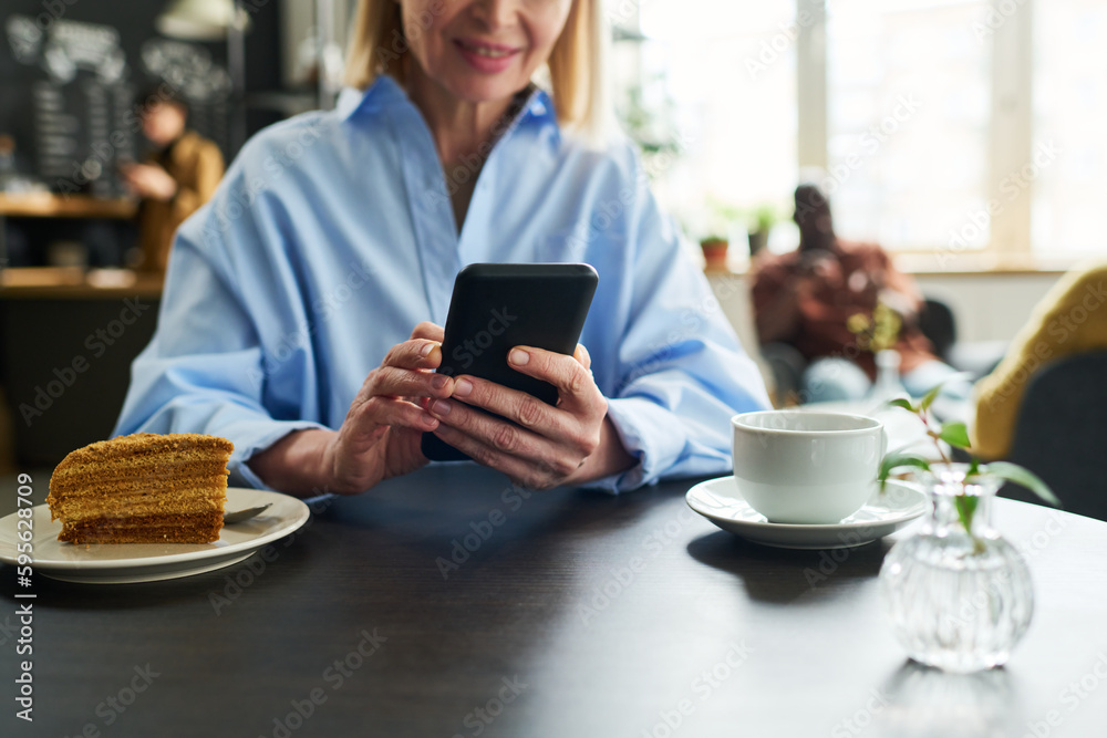 Close-up of mature female consumer using smartphone by table served with cup of tea and piece of cake while having breakfast in cafe