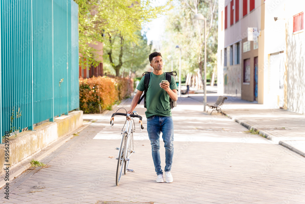 Fototapeta premium Cheerful ethnic man with bicycle walking with backpack delivering food