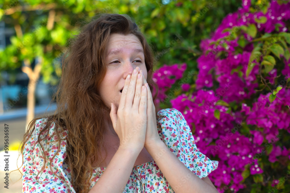 Portrait of beautiful young allergic woman is suffering from pollen allergy or cold on natural flower flowering tree background at spring or sunny summer day sneezes, blowing her runny nose rubs eyes