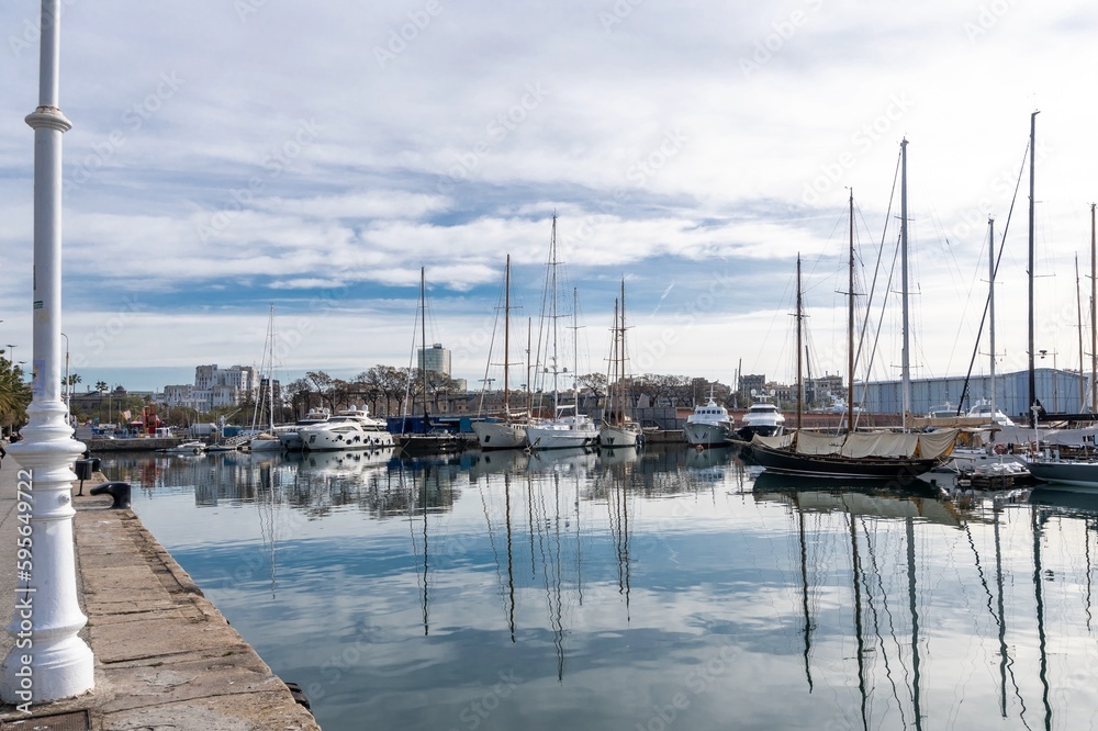Barcelona, Spain-April 12, 2023. A tranquil scene of a sailboats gliding in the harbor with reflections on the still water, surrounded by Barcelona port vell and its nautical vessels