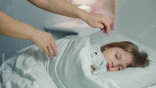 Cute funny little girl with brown hair sleeping in the white bed with favorite fluffy toy bunny. Woman hand covers with blanket caressing lying daughter. Good night. Bedtime rest. Carefree childhood