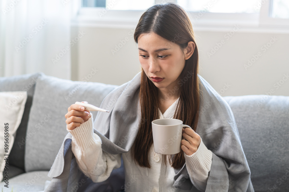 Sick, influenza asian young woman, girl have a fever, hand holding thermometer for check measure body temperature and drink hot water, illness while sitting rest on sofa at home. Health care concept. Sick, influenza asian young woman, girl have a fever, hand holding thermometer for check measure body temperature and drink hot water, illness while sitting rest on sofa at home. Health care concept.