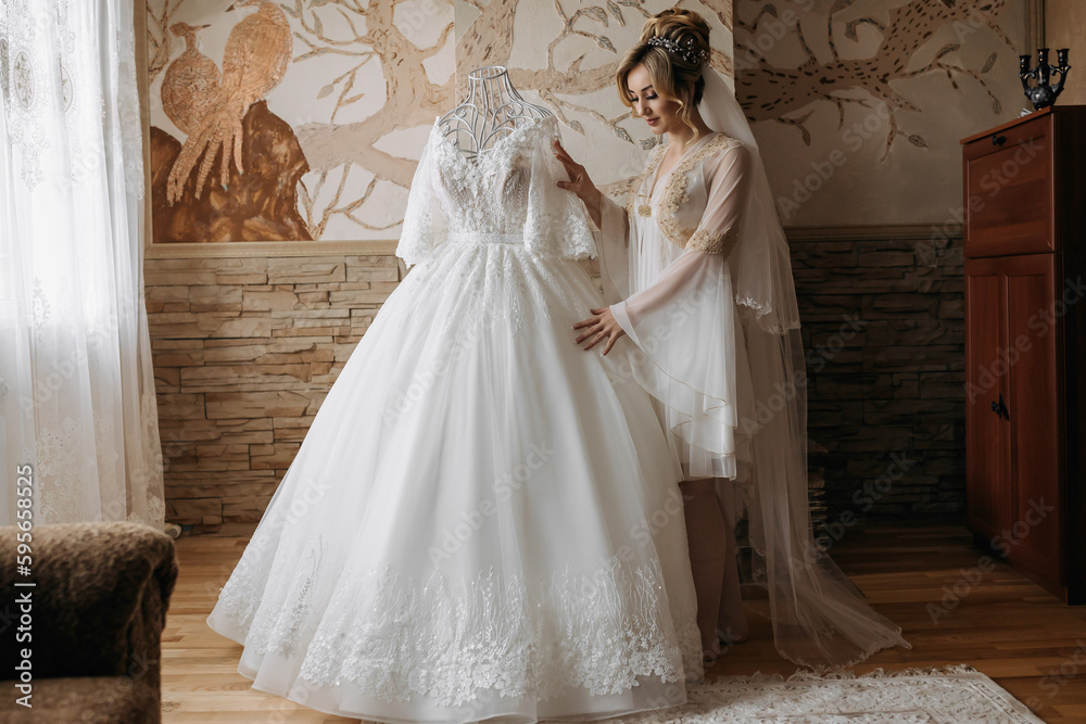 Fototapeta premium Blonde Bride in a robe, posing for the camera looking at her wedding dress. Elegant hairstyle. Diadem. Beautiful makeup. Morning of the bride