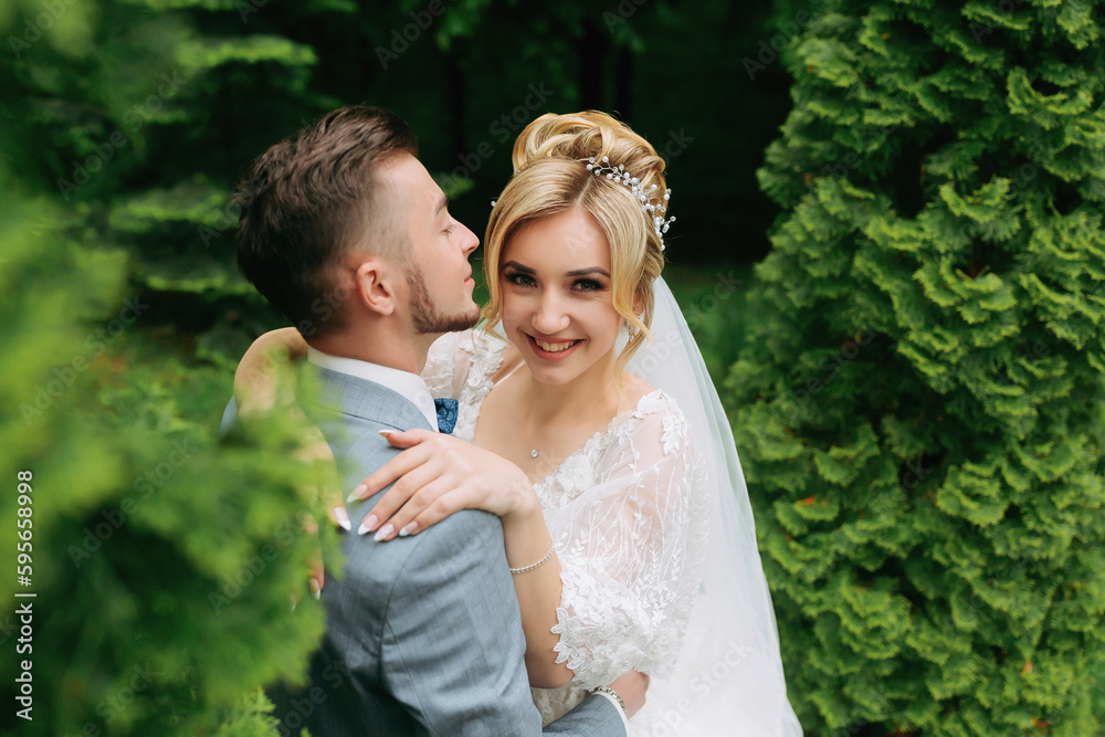 Portrait of the bride and groom standing on the background of green trees, embracing. The bride smiles sincerely. Stylish groom. Fashion and style. Beautiful bride
