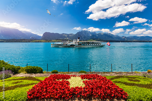 Fototapeta Naklejka Na Ścianę i Meble -  Flowerbed of the Swiss flag with boat cruise on the Thun lake and Alps mountains, Oberhofen, Switzerland. Swiss flag made of flowers and passenger cruise boat, Lake Thun, Switzerland.