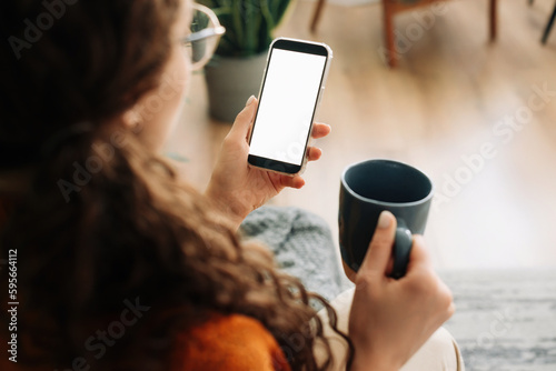 Over-shoulder view of woman holding coffee and smartphone with white mock-up cellular screen in hands using mobile apps on a cell phone. Cellphone display mock-up for advertising concept.