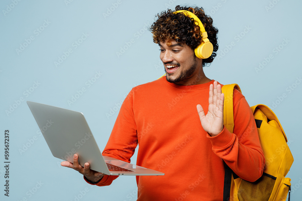 Young teen Indian boy student wearing casual clothes backpack bag headphones listen music hold laptop pc computer talk speak isolated on plain blue background. High school university college concept.