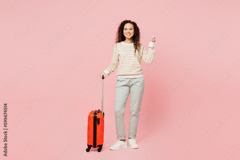 Traveler woman wear casual clothes hold suitcase point finger overhead isolated on plain pastel pink background. Tourist travel abroad in free spare time rest getaway. Air flight trip journey concept.