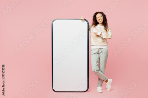 Full body smiling young woman wearing light casual clothes point index finger on big huge blank screen mobile cell phone smartphone with area isolated on plain pastel pink background studio portrait.